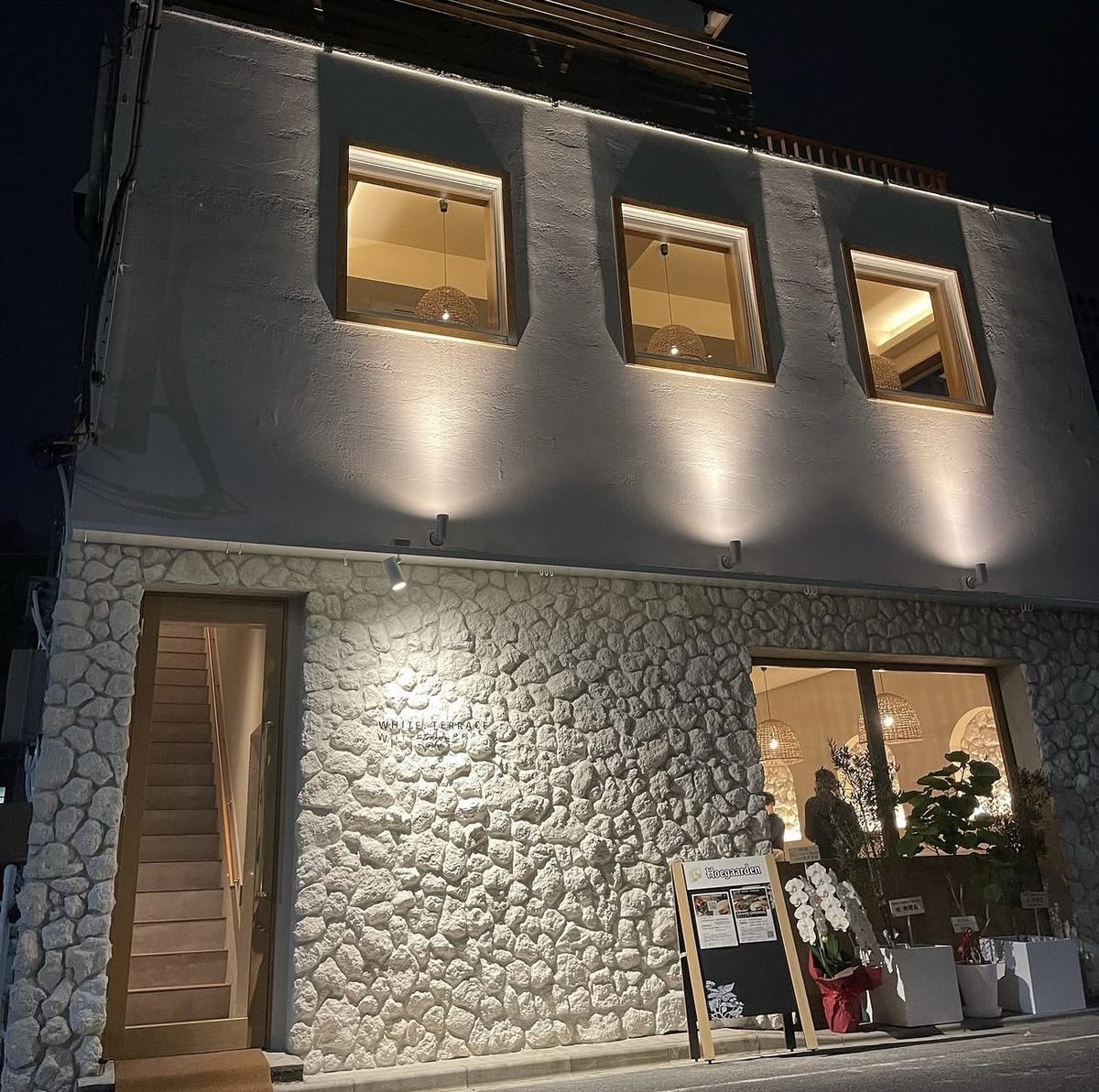 夜の外観。1階は白い石積み調の壁、2階は白い塗り壁で、四角い窓が三つ並ぶ。壁に沿って照明が当たり、入口の奥に階段が見える
